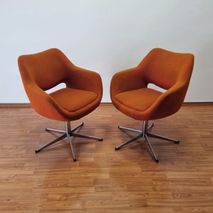 May include: Two orange swivel chairs with a mid-century modern design. The chairs have a rounded seat and back with a cut-out detail. They sit on a silver, star-shaped base. The chairs are placed on a wood-grain floor.