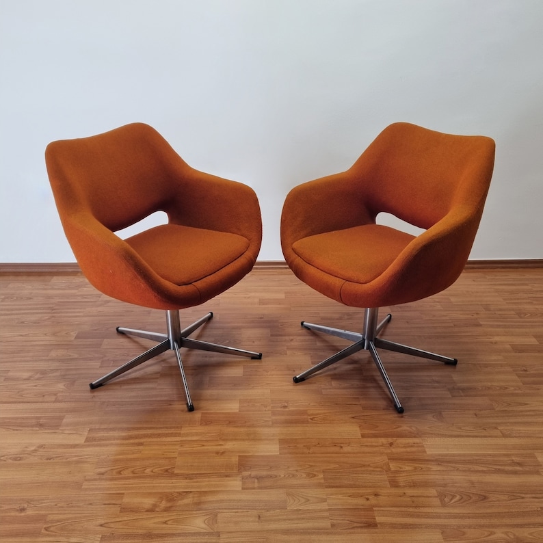 May include: Two orange swivel chairs with a mid-century modern design. The chairs have a rounded seat and back with a cut-out detail. They sit on a silver, star-shaped base. The chairs are placed on a wood-grain floor.