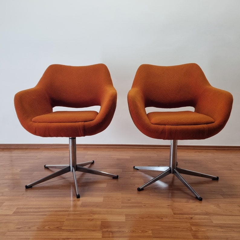 May include: Two orange swivel chairs with a mid-century modern design. The chairs have a rounded seat and back with a cut-out detail. They sit on a silver, star-shaped base. The chairs are placed on a wood floor.