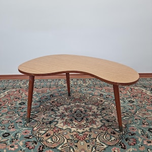 Op de afbeelding: Een moderne salontafel in nierenvorm uit het midden van de eeuw met een tafelblad van houtlaminaat en vier slanke houten poten. De tafel staat op een tapijt met een patroon.