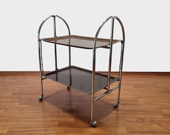 May include: A chrome serving cart with two trays. The top tray is brown and the bottom tray is black. The cart has four wheels and is on a wooden floor.