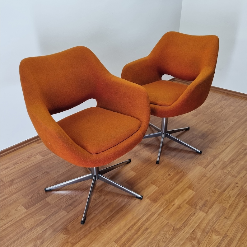 May include: Two orange swivel chairs with a mid-century modern design. The chairs have a curved back and armrests, and a seat cushion. They sit on a silver, star-shaped base. The chairs are placed on a wooden floor.