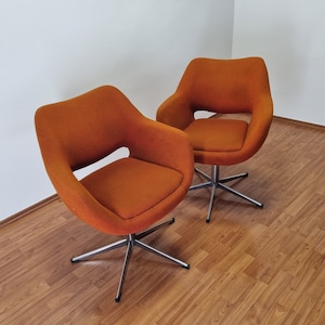 Può includere: Due poltrone girevoli arancioni dal design moderno della metà del secolo. Le sedie hanno schienale e braccioli curvi, con un cuscino di seduta. La base è in metallo argentato con cinque gambe.