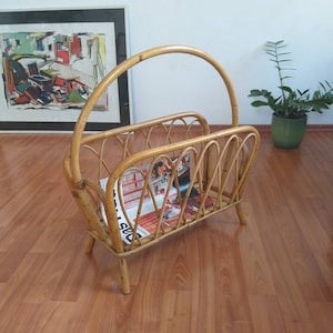 Puede incluir: Un revistero de bambú natural tejido con un asa superior curva. El revistero contiene una revista con una imagen colorida de una mujer en la portada.