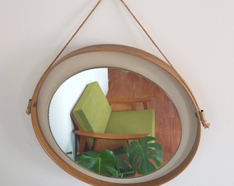 May include: A round wooden framed mirror with a rope hanger. The mirror reflects a green armchair and a potted plant.