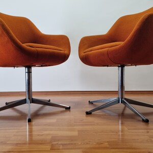 May include: Two mid-century modern swivel chairs. The chairs have a rounded, orange fabric seat and back. They are mounted on a silver, star-shaped base. The chairs are set against a white wall and a wooden floor.