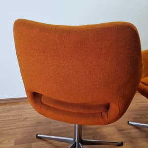 May include: An orange, mid-century modern style chair with a curved back and a swivel base. The chair has a textured fabric and a stainless steel base. The chair is set against a white wall and a wooden floor.