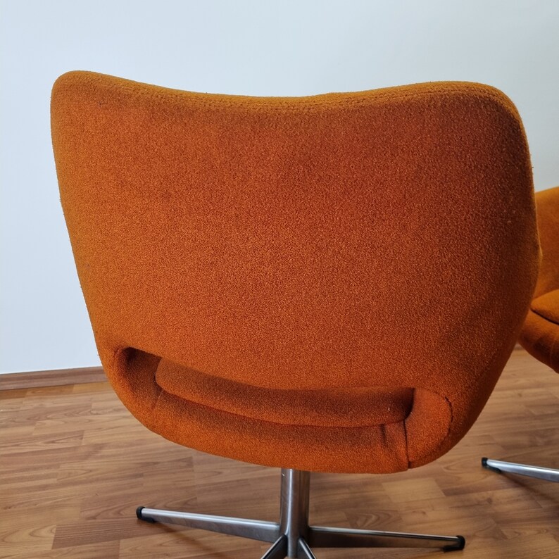 May include: An orange, mid-century modern style chair with a curved back and a swivel base. The chair has a textured fabric and a stainless steel base. The chair is set against a white wall and a wooden floor.