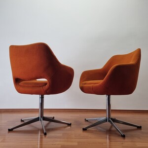 May include: Two orange swivel chairs with a mid-century modern design. The chairs have a curved back and armrests, with a cut-out detail on one side. They sit on a silver, star-shaped base.