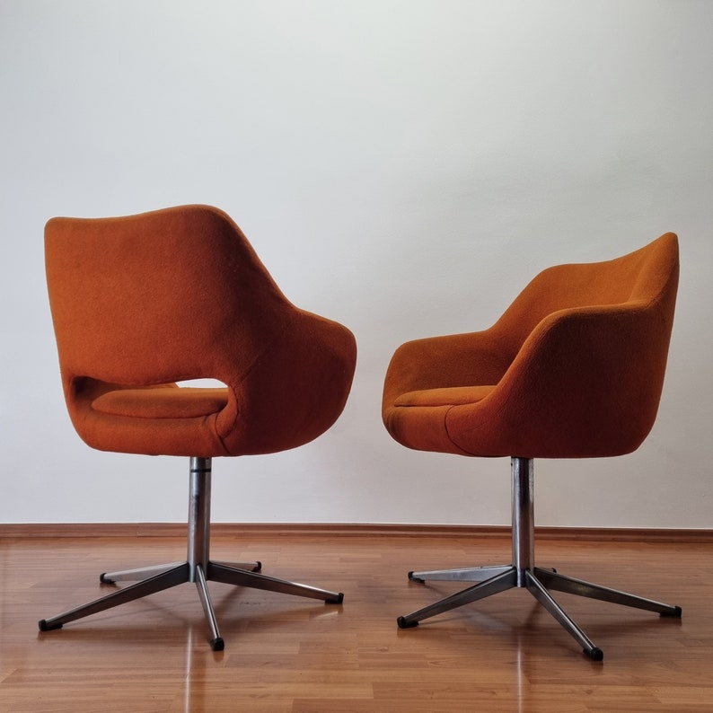 May include: Two orange swivel chairs with a mid-century modern design. The chairs have a curved back and armrests, with a cut-out detail on one side. They sit on a silver, star-shaped base.