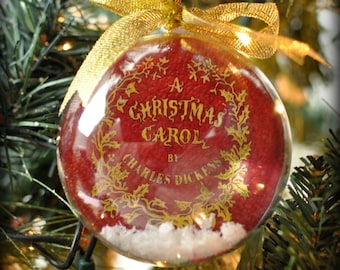 May include: A clear Christmas ornament with the text "A Christmas Carol by Charles Dickens" in gold lettering. The ornament has a red interior with a snow-like base and is topped with a gold ribbon, hanging on a Christmas tree.