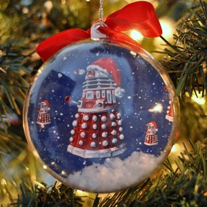 May include: A clear Christmas ornament with a red ribbon bow, featuring a red and white Dalek wearing a Santa hat against a snowy blue background. The ornament is hanging on a Christmas tree.