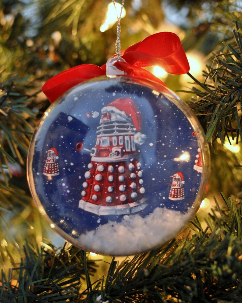 May include: A clear Christmas ornament with a red ribbon bow, featuring a red and white Dalek wearing a Santa hat against a snowy blue background. The ornament is hanging on a Christmas tree.