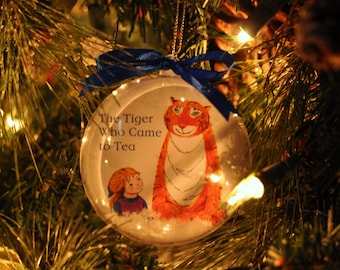 May include: Clear glass Christmas ornament featuring an illustration of a tiger and a girl with the text "The Tiger Who Came to Tea." The ornament is decorated with a blue ribbon and hangs on a Christmas tree.