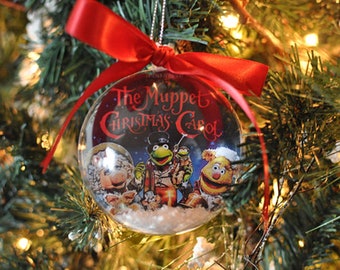 May include: A clear Christmas ornament featuring characters from "The Muppet Christmas Carol" movie. The ornament has a red ribbon and is hanging on a Christmas tree. The text "The Muppet Christmas Carol" is visible on the ornament.