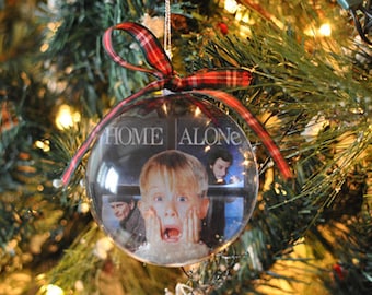 May include: Clear glass Christmas ornament featuring a scene from the movie "Home Alone." The ornament has a red and green plaid ribbon for hanging. The ornament is hanging on a Christmas tree with green needles and lights.