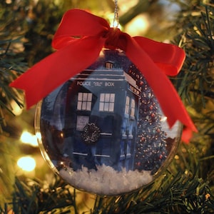 May include: A clear Christmas ornament with a blue police box design inside, the words "POLICE CALL BOX" are visible. The ornament is decorated with artificial snow and a red ribbon. The ornament hangs on a Christmas tree.