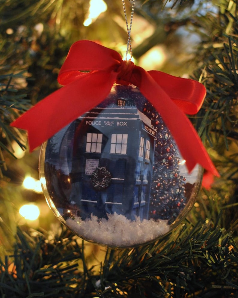 May include: A clear Christmas ornament with a blue police box design inside, the words "POLICE CALL BOX" are visible. The ornament is decorated with artificial snow and a red ribbon. The ornament hangs on a Christmas tree.