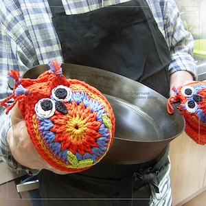 Puede incluir: Un par de agarraderas de olla de búho de ganchillo en rojo, naranja, amarillo, azul y verde. Las agarraderas tienen ojos negros y pupilas blancas. Las agarraderas están siendo sostenidas por una persona que lleva una camisa a cuadros y un delantal negro.