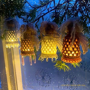 May include: Four handmade crochet angel decorations with internal lights, suspended from a dark green branch. The angels feature white yarn hair and wings, emitting a warm glow. The text "cottonheartland + bright lotus bidder design" is visible.