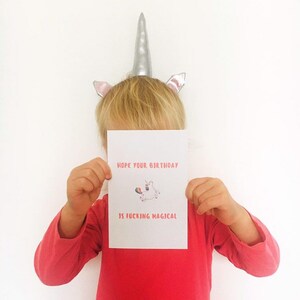 Puede incluir: Una tarjeta blanca con texto rosa que dice "Espero que tu cumplea&ntilde;os sea jodidamente m&aacute;gico." Un unicornio de dibujos animados con una cola de arco&iacute;ris est&aacute; ilustrado en la tarjeta. Un ni&ntilde;o con una diadema de unicornio plateado est&aacute; sosteniendo la tarjeta frente a su cara.