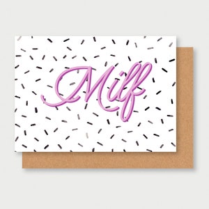 Peut inclure: Une carte blanche avec un texte "Milf" en rose en 3D sur un fond de confettis noirs et gris. La carte a une enveloppe brune.