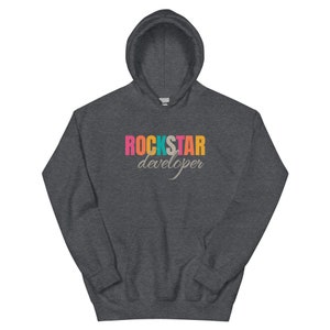 Op de afbeelding: Donkergrijze hoodie met een trekkoord en een voorzak. De voorkant toont de tekst "ROCKSTAR developer" in een retro-lettertype. Het woord "ROCKSTAR" is roze, turquoise, oranje en geel.