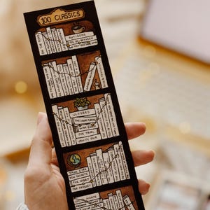 May include: A bookmark with a hand-drawn bookshelf design and the text "100 Classics." The bookmark shows various book titles, including "Pride & Prejudice" and "The Great Gatsby," with a brown background and white book spines.
