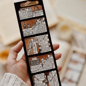 May include: A black bookmark with a hand-drawn illustration of books, a typewriter, and candles. The bookmark is labelled "100 CLASSICS" and lists book titles such as "Hamlet" and "Moby Dick". The bookmark is held by a person.