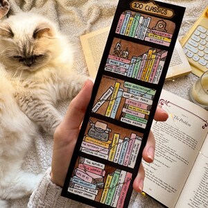 May include: A black bookmark with a hand-drawn bookshelf design and the text "100 CLASSICS." The bookmark shows miniature book titles such as "1984" and "Frankenstein." The image is held against a soft, textured background.