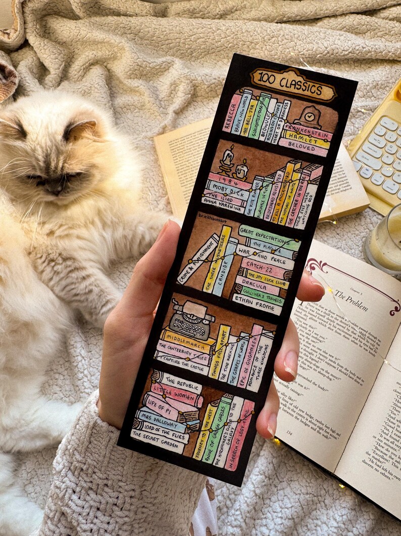 May include: A black bookmark with a hand-drawn bookshelf design and the text "100 CLASSICS." The bookmark shows miniature book titles such as "1984" and "Frankenstein." The image is held against a soft, textured background.