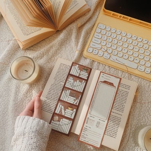 May include: An open book with a decorative bookmark featuring a bookshelf design. A second bookmark with a reading log is visible. A lit candle and a yellow tablet with a keyboard are also in the image. The scene is set on a textured, off-white surface.