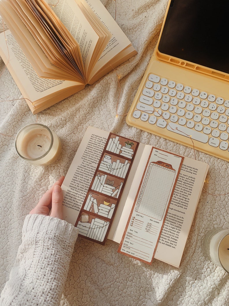 May include: An open book with a decorative bookmark featuring a bookshelf design. A second bookmark with a reading log is visible. A lit candle and a yellow tablet with a keyboard are also in the image. The scene is set on a textured, off-white surface.