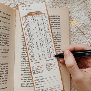 May include: A bookmark with a reading tracker for the year 2021, with a goal of 25 books. The bookmark is brown with a grid for marking days and book details. A hand is holding a black marker, marking the tracker.