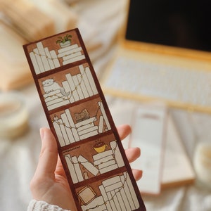 May include: A brown bookmark with a bookshelf design. The bookmark features illustrations of books, a cat, a plant, candles, and a cup of coffee. The text "britishbookart" is visible on the bookmark.