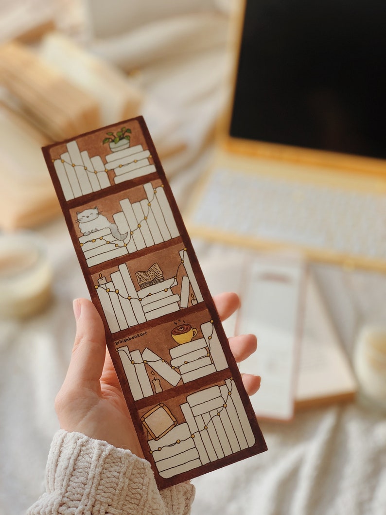 May include: A brown bookmark with a bookshelf design. The bookmark features illustrations of books, a cat, a plant, candles, and a cup of coffee. The text "britishbookart" is visible on the bookmark.