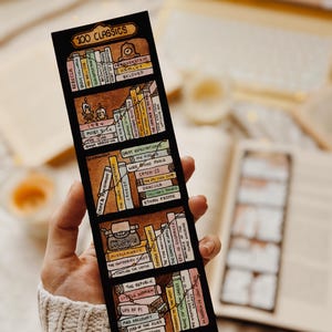 May include: A black bookmark with a hand-drawn bookshelf design and the text "100 CLASSICS." The bookmark displays miniature book spines with titles such as "Rebecca," "Hamlet," and "Great Expectations." The design includes a typewriter and candles.