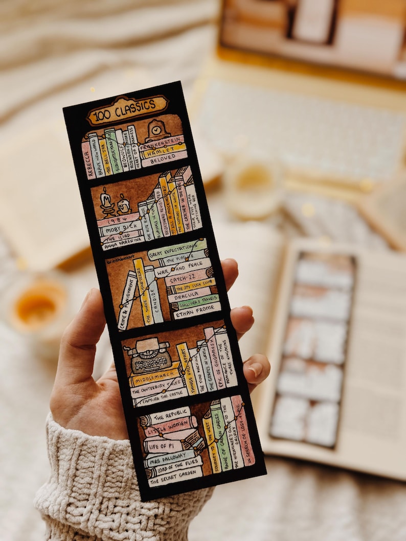 May include: A black bookmark with a hand-drawn bookshelf design and the text "100 CLASSICS." The bookmark displays miniature book spines with titles such as "Rebecca," "Hamlet," and "Great Expectations." The design includes a typewriter and candles.