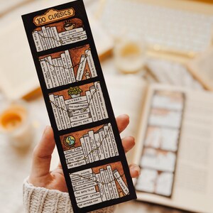 May include: A black bookmark with the text "100 CLASSICS" and illustrations of book spines. The bookmark features titles like "Jane Eyre," "The Hobbit," and "Ulysses." The design includes a brown and beige color scheme, with a hand holding the bookmark.