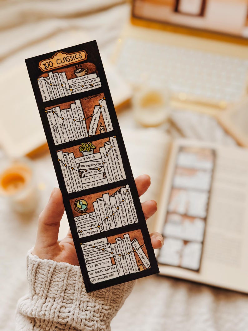 May include: A black bookmark with the text "100 CLASSICS" and illustrations of book spines. The bookmark features titles like "Jane Eyre," "The Hobbit," and "Ulysses." The design includes a brown and beige color scheme, with a hand holding the bookmark.