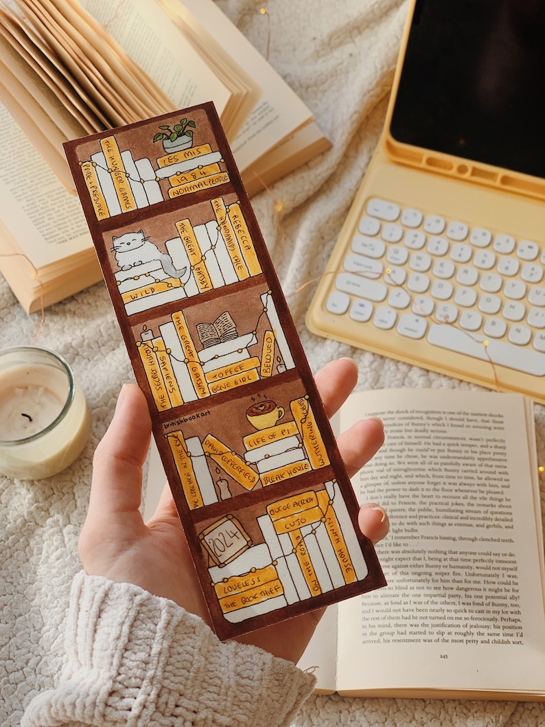 May include: A handmade bookmark featuring a bookshelf design with book titles like "Les Mis" and "1984." The bookmark is brown with white and yellow accents, and includes a small cat illustration. A small plant and a cup of coffee are also depicted.