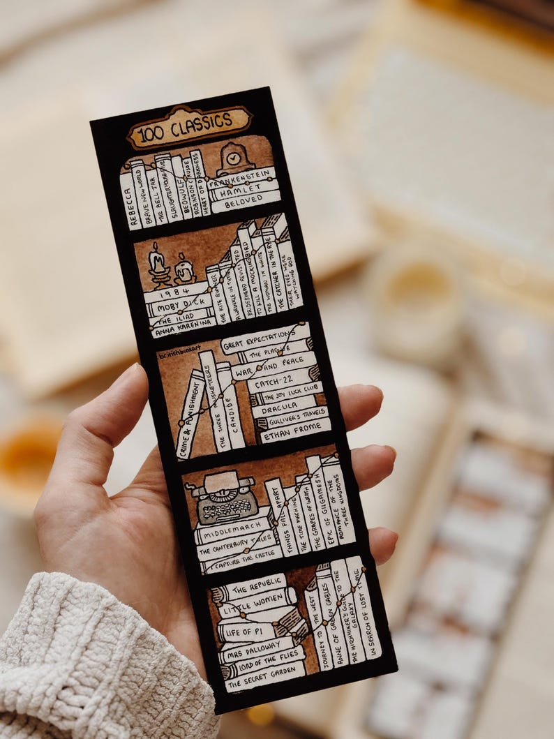 May include: A black bookmark with a hand-drawn illustration of books, a typewriter, and candles. The bookmark is labelled "100 CLASSICS" and lists book titles such as "Hamlet" and "Moby Dick". The bookmark is held by a person.