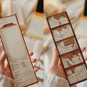 May include: Two brown paper bookmarks. One features a reading tracker with a grid, the text "DAYS I READ", and a star tally. The other bookmark depicts a bookshelf with books, a cat, and a cup of coffee. Both bookmarks are held by a person.