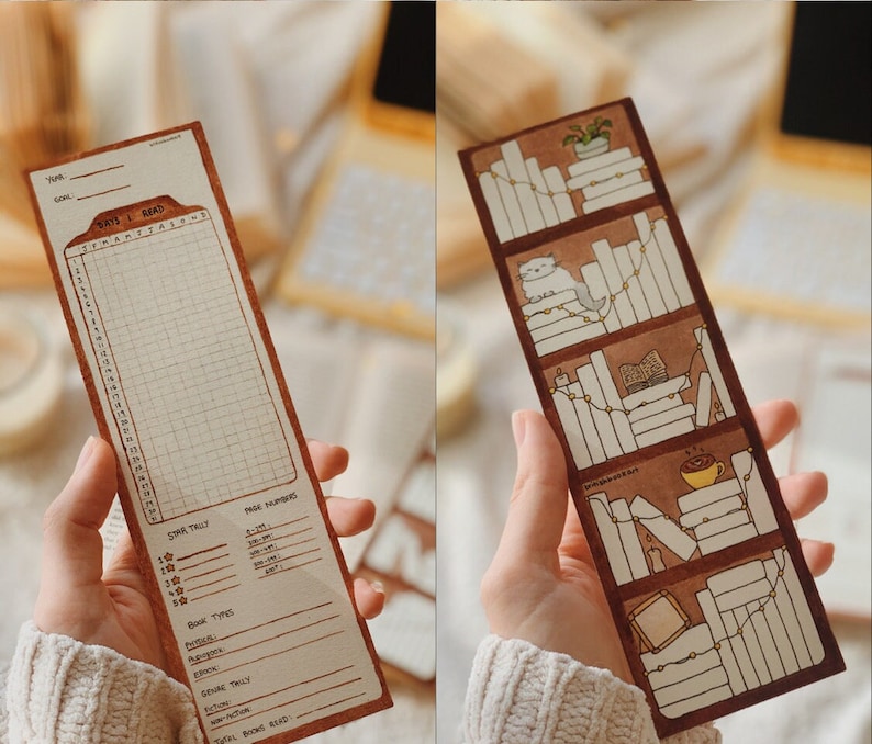 May include: Two brown paper bookmarks. One features a reading tracker with a grid, the text "DAYS I READ", and a star tally. The other bookmark depicts a bookshelf with books, a cat, and a cup of coffee. Both bookmarks are held by a person.