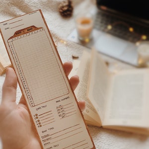May include: A tan bookmark with a reading tracker grid, star tally, and sections for book types and genres. The bookmark has the text "DAYS I READ" and "STAR TALLY". The bookmark is held in a hand, with a book and other items in the background.