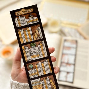 May include: A bookmark with a hand-drawn illustration of a bookshelf, labelled "100 Classics." The design features various book titles in brown and yellow, with a decorative line connecting the books. The bookmark is held by a person, showcasing the design.