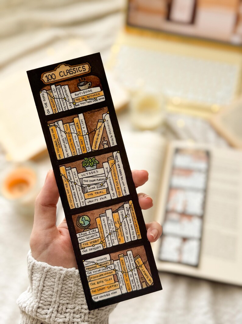May include: A bookmark with a hand-drawn illustration of a bookshelf, labelled "100 Classics." The design features various book titles in brown and yellow, with a decorative line connecting the books. The bookmark is held by a person, showcasing the design.