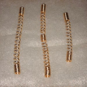 May include: Three copper-colored spiral springs of varying lengths. The springs are tightly coiled and have a shiny, metallic finish. They are arranged against a white, textured background, showcasing their form and detail.