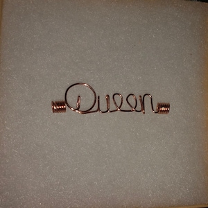 Puede incluir: Un accesorio para el cabello de alambre de color cobre que deletrea la palabra "Queen". La palabra está en una fuente cursiva, con acentos de alambre enrollado en cada extremo. El accesorio se muestra sobre una superficie blanca.