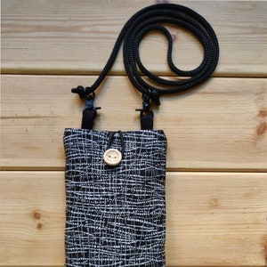 Pode incluir: Uma bolsa para telefone com padr&atilde;o preto e branco com uma al&ccedil;a ajust&aacute;vel preta. A bolsa tem um pequeno fecho de bot&atilde;o de madeira.